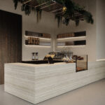 Zijaanzicht van een horecagelegenheid met bar uitgewerkt in plaatmateriaal FD01 Taranta van Cleaf, welke de beleving van travertin heeft. De wandafwerking uitgewerkt in een zandkleurig decor FD10 Taranta van Cleaf en donkerbruine tint van LN55 Maloja, een decor dat is geïnspireerd op mat gelakt notenhout.