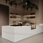Zijaanzicht van een horecagelegenheid met een bar uitgewerkt in het solid surface decor DM5048 Starstruck van Durasein, welke de luxe uitstraling van marmer op een warm witte basis biedt. De wandafwerking uitgewerkt in de FD29 Taranta van Cleaf, welke de beleving van zandsteen geeft, en een deur in de FD14 Taranta van Cleaf, een bruine steentextuur.